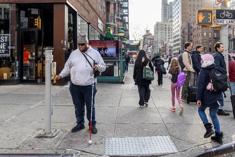 A person who uses a white cane presses an APS button while waiting at a corner in New York City.