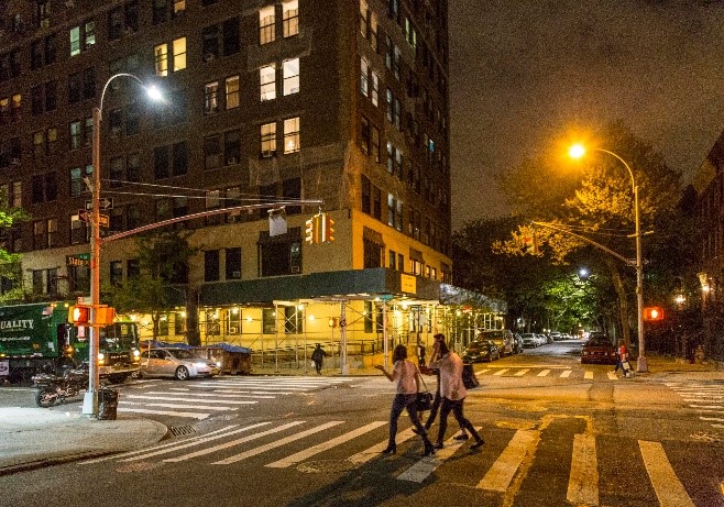 At night people walk over a crosswalk in NYC, illuminated by tall streetlights.