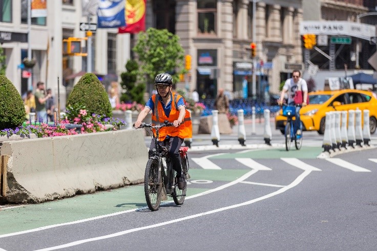 A person wearing a helmet and orange safety vest rides an e-bike on a bike lane in Manhattan.