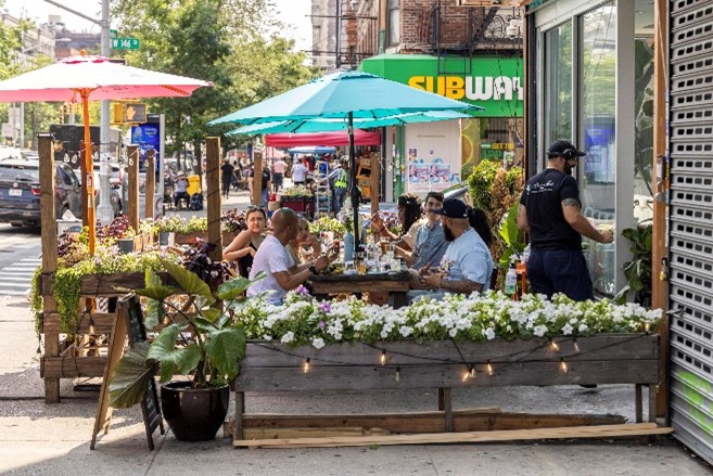 People sit at a restaurant with tables and umbrellas set up on a wide sidewalk on a sunny day.