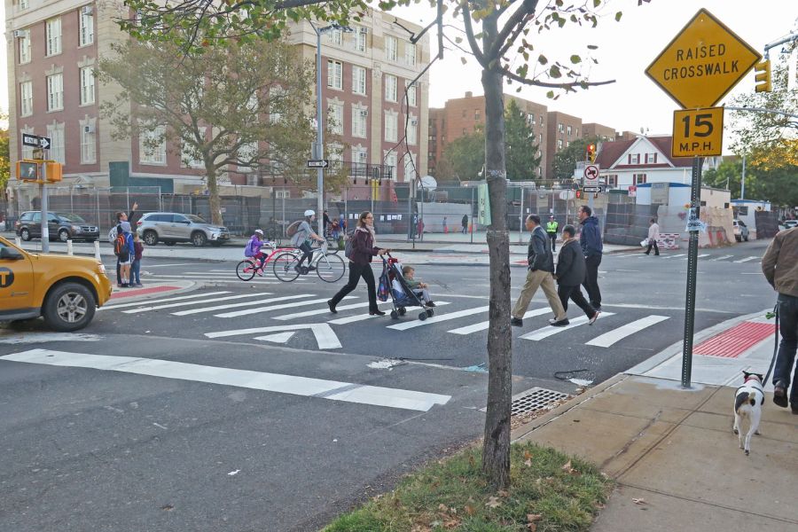 A group of three adults, a person pushing a stroller walk in a crosswalk on a raised speed hump.
