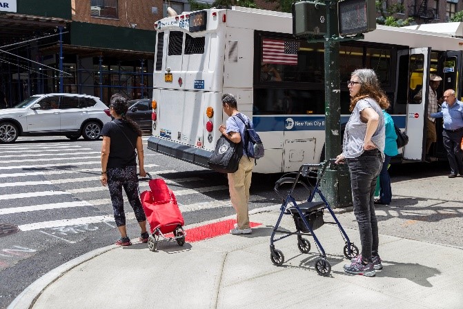 Three adults wait at a corner in New York City, one person pulls a small red carriage and another person stands with a walker.