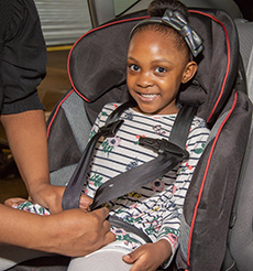 A child smiles at the camera as she’s properly buckled into a car seat.