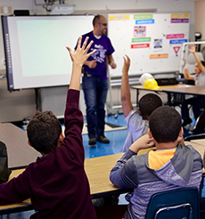 An educator stands in front of a classroom as the students sitting around him raise their hands.