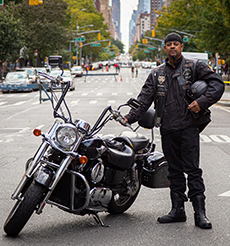 A man holding a helmet stands next to a parked motorcycle on a street closed to traffic.