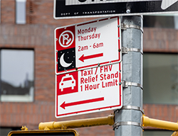 Two parking regulation signs on a pole. One designates the space as a Taxi / F H V Relief Stand and the other shows street sweeping times.