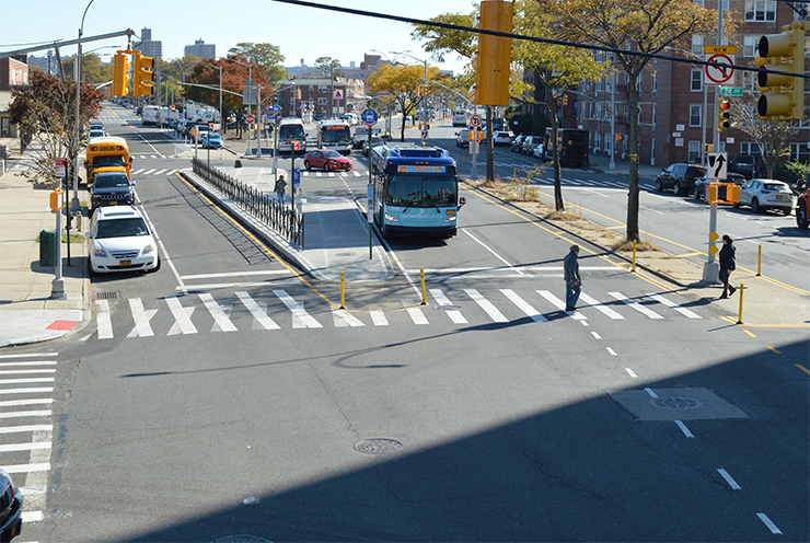 Two pedestrians cross Cropsey Avenue at 24th Avenue with a painted median tip and a bus boarding island reducing pedestrian crossing distance and exposure.