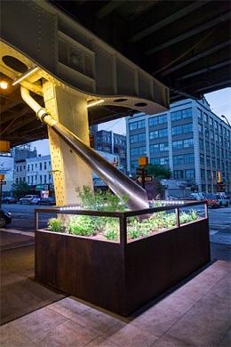 A brightly light space below an elevated roadway featured a stylist drainpipe leading to a tall, illuminated planter with green plants.