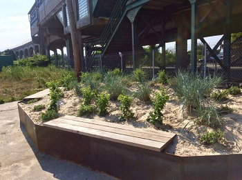 On a sunny day, a large planter is filled with green shrubs and benches, located at the base of an outdoor staircase beside an elevated train.