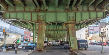 A rusting structure supporting an elevated roadway, with cars parked below it.