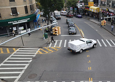 A car turns left around a complete hardened centerline treatment, helping the driver to see a pedestrian walking in the crosswalk. Location is Grand Ave & West Burnside Ave in the Bronx, NY.