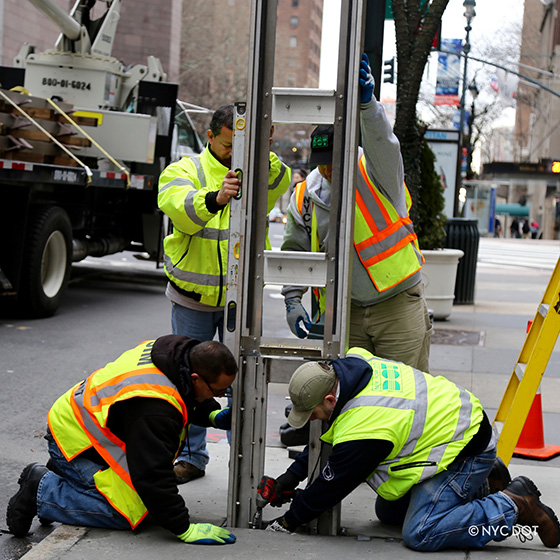 Four DOT crew members in bright safety jacket install a tall metal frame on a sidewalk, near the curb.