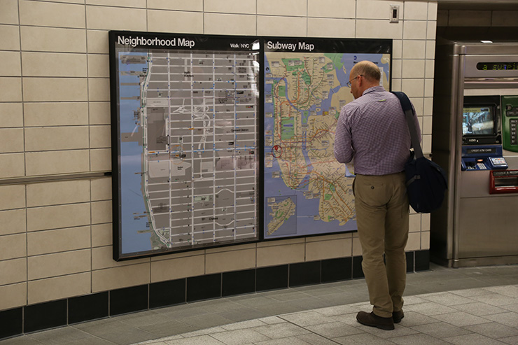 A man stands in a NYC subway station and looks at two large map posters hanging on the station wall