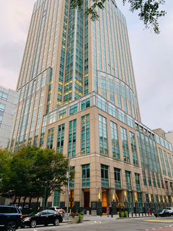 12 Metrotech Center building. The color of the building is beige with blue windows. There are trees next to the building and cars are driving by.