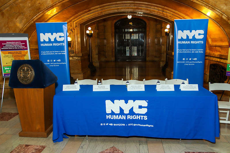 A formal event setup inside an ornate, arched hall at New York City Hall, featuring a long table draped in a blue NYC Human Rights tablecloth with name placards and water bottles arranged for a panel discussion. Two vertical NYC Human Rights banners stand behind the table, and a wooden podium with the City of New York seal is positioned to the left. The space is warmly lit, with marble floors and a gated doorway centered in the background.