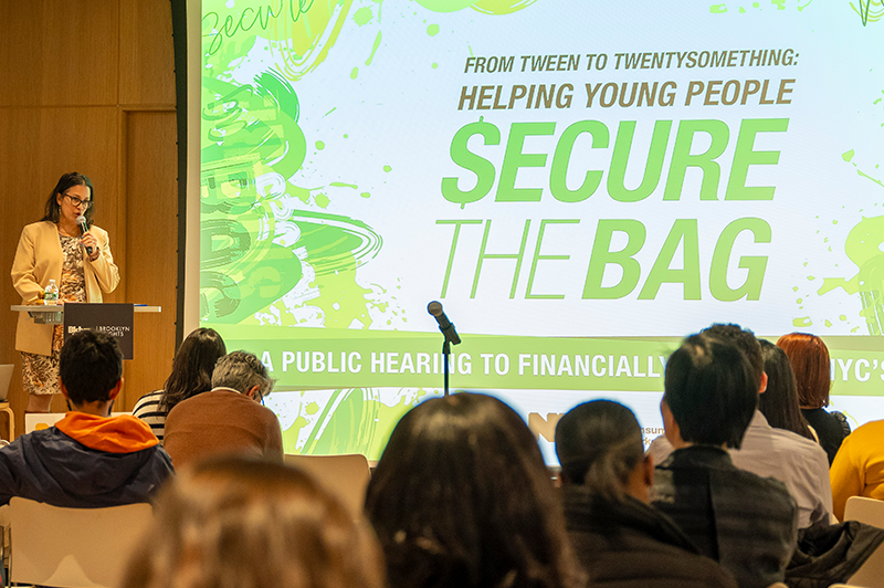 Commissioner Mayuga at podium providing opening remarks during public hearing event, From Tween to TwentySomething: Helping Young People Secure the Bag
                                           