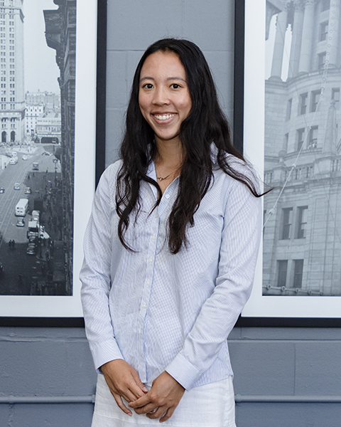 Woman smiling, wearing a button down light blue shirt with long sleeves.