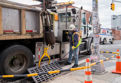 a dep crew member stands next to a truck to control the mechanism that's lifting the grates off of the catch basin in the street.