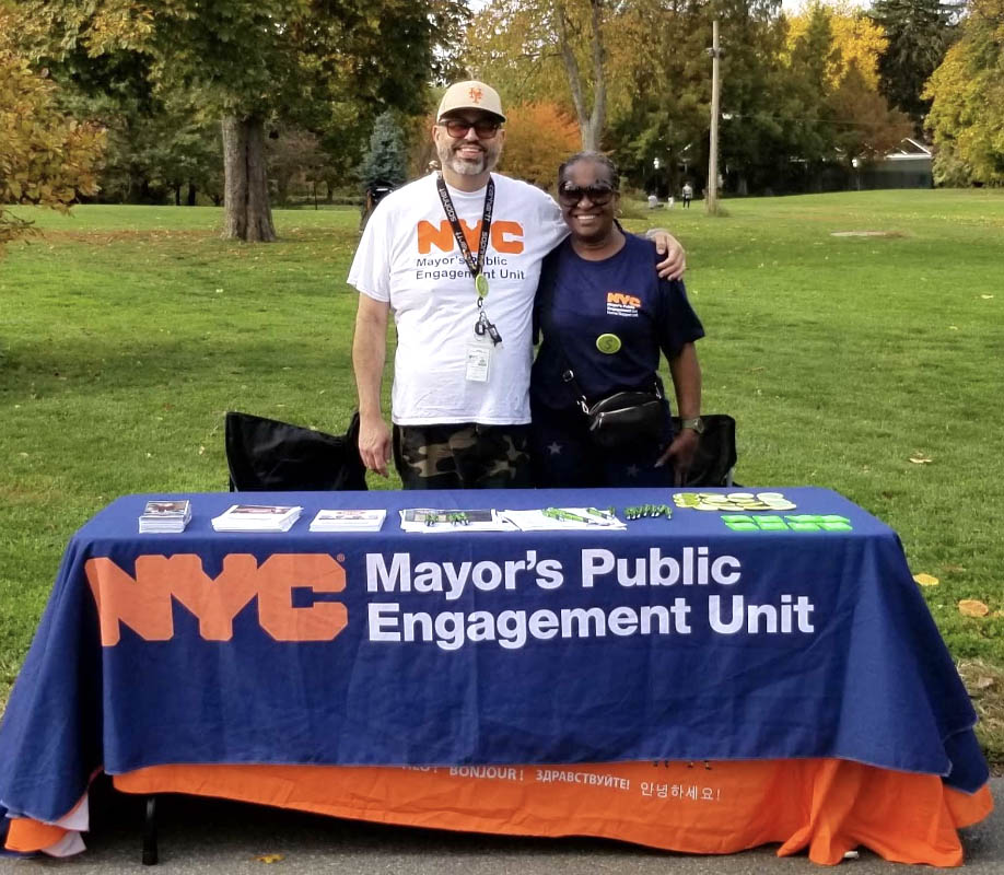 Two PEU staff wearing PEU shirts hug and smile while standing behind table that holds flyer and table cloth that reads NYC Mayor's Public Engagement Unit.