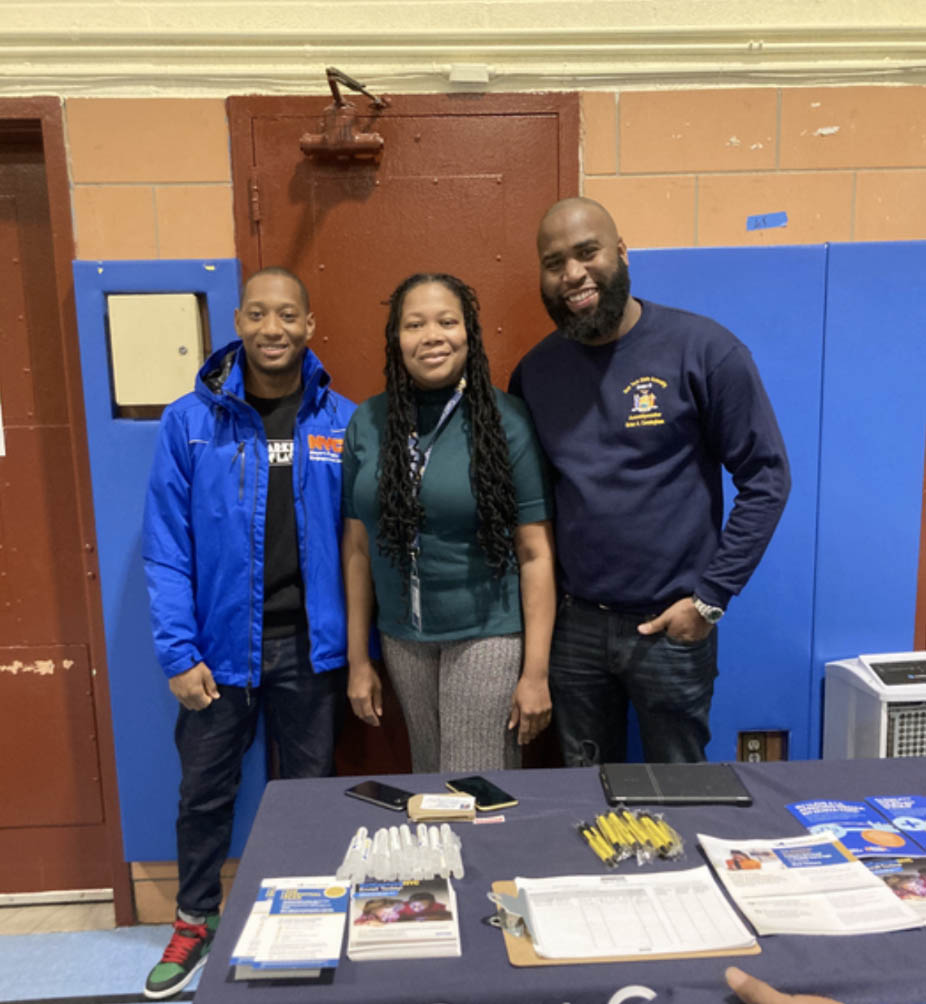 Two PEU staff members stand by a PEU table with Assembly Member Cunningham inside what appears to be a school gym.