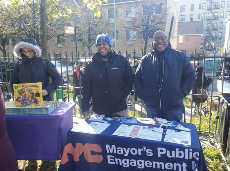 Two PEU staff stand behind a table smiling that hold flyers and table cloth that reads NYC Mayor's Public Engagement.