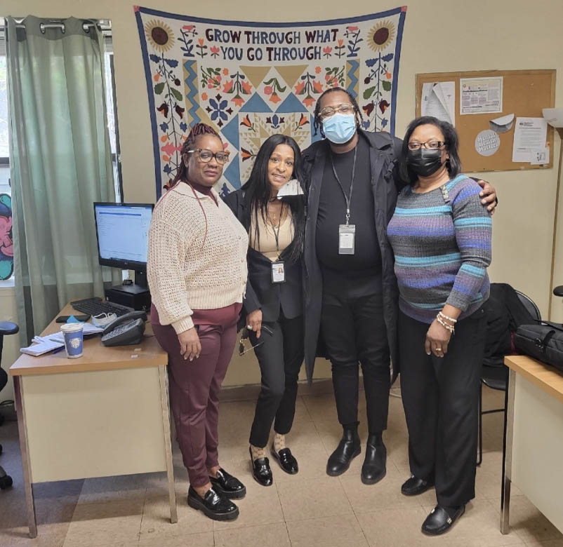 4 PEU Specialists stand together next two a couple of desks in a small office. Behind them is a quilt on the wall with text that reads Grow through what you go through.