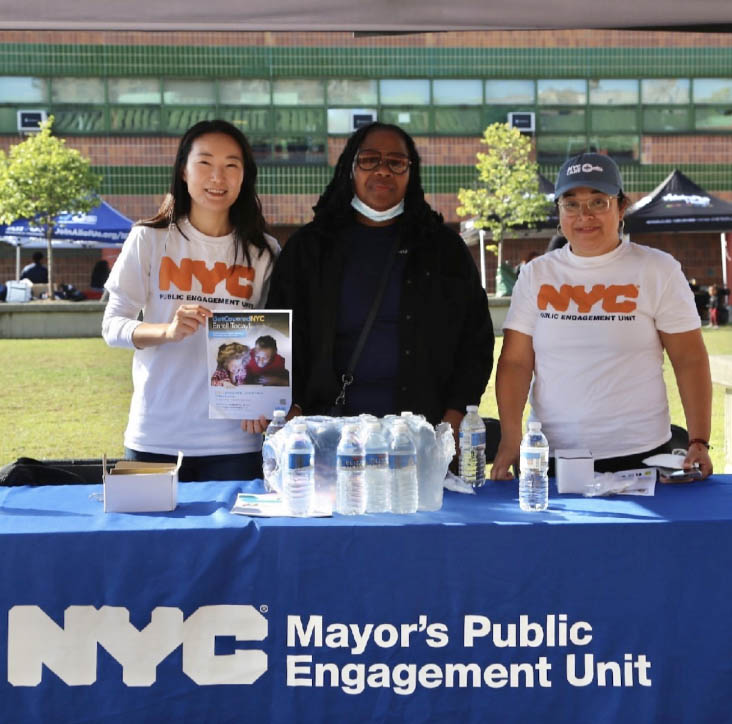 Three Get Covered N Y C Specialists stand behind the PEU table. They wear PEU shirts and one holds a flyer up that has information about GetCoveredNYC on it.