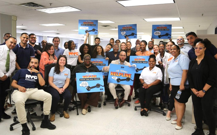 Big group of Tenant Support staff in a PEU office, smiling together, with some holding up signs about the Tenant Helpline