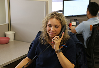 Woman sits in chair wearing a navy blue PEU polo while on a landline phone smiling