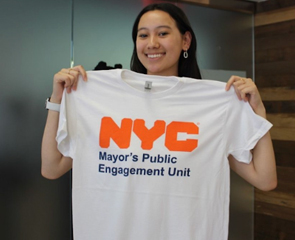 Young woman smiles as she holds up a white T-Shirt with the logo that reads NYC Mayor's Public Engagement Unit