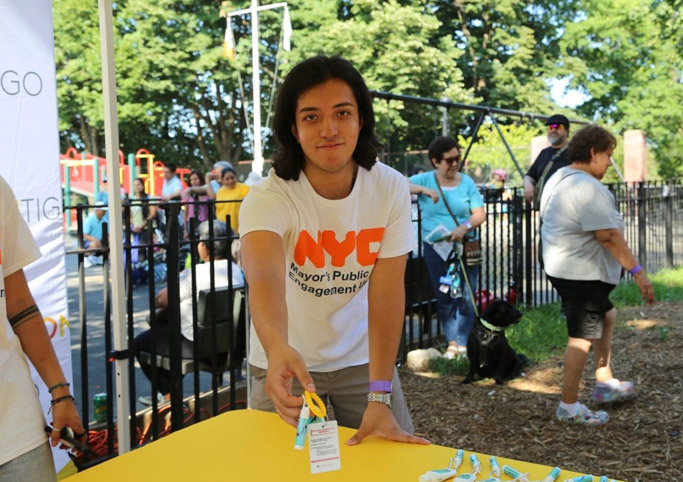 Young man wearing a NYC Mayor's Public Engagement Unit T-Shirt stands behind a table and smiles as he is handing over a key chain with water safety tips