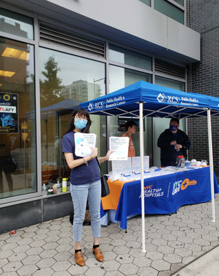 Woman stands in front of NYC Health and Hospitals booth as she holds up two flyers.