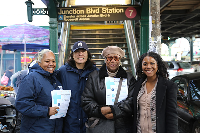 Four women stand in front of the Junction Blvd 7 train station smiling. Two of them hold flyers