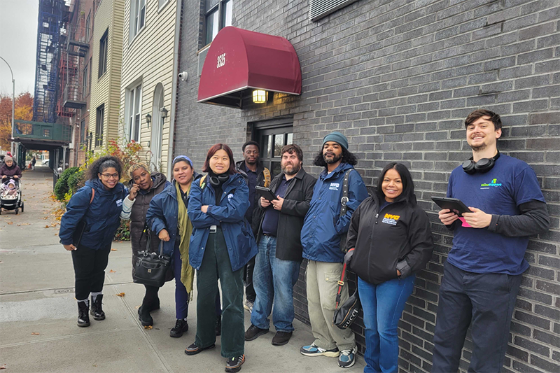 Nine staff members from PEU's Housing Support Unit pose in Public Engagement Unit gear outside of a large apartment building on an NYC sidewalk