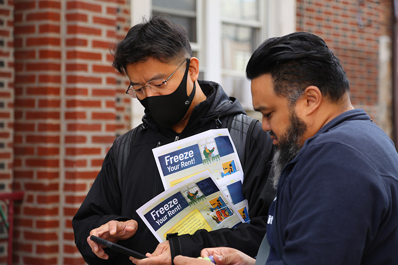 Two men stand outside a building holding flyers titled "Freeze your Rent" while looking at their work cellphones