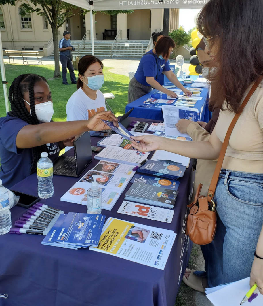 Table with flyers and markers. PEU staff sit at one side of the table while wearing masks and PEU tee shirts while students stand on the other side of the table. one PEU staff is handing a flyer to a student