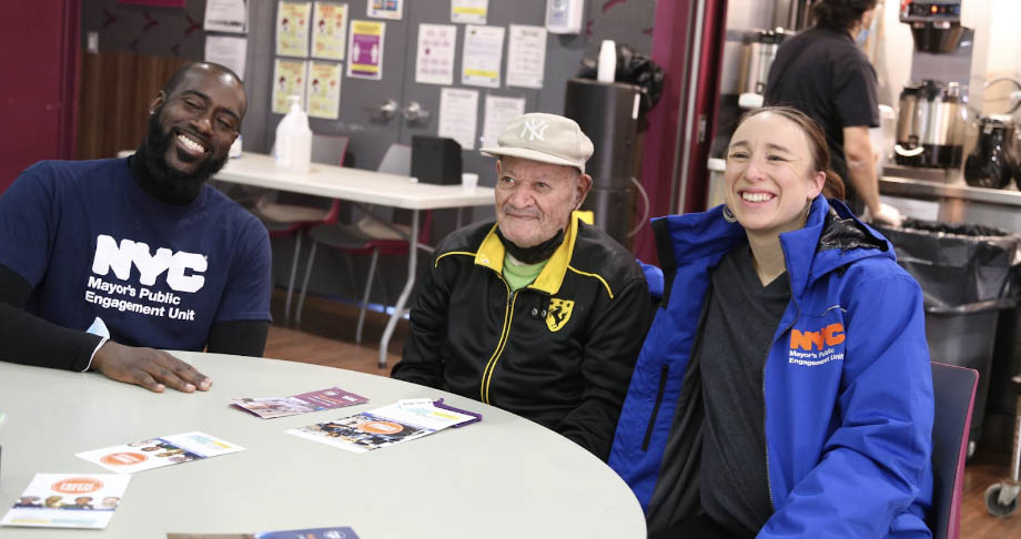 Chris Wagner and Adrienne Lever sit next to an older adult wearing a hat while they smile at the camera and wear PEU mech