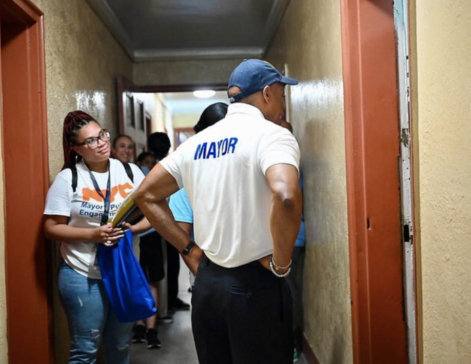 PEU staff and mayor Eric Adams ,who is wearing a white shirt that says mayor and a blue hat, stand outside an apartment door that is open.