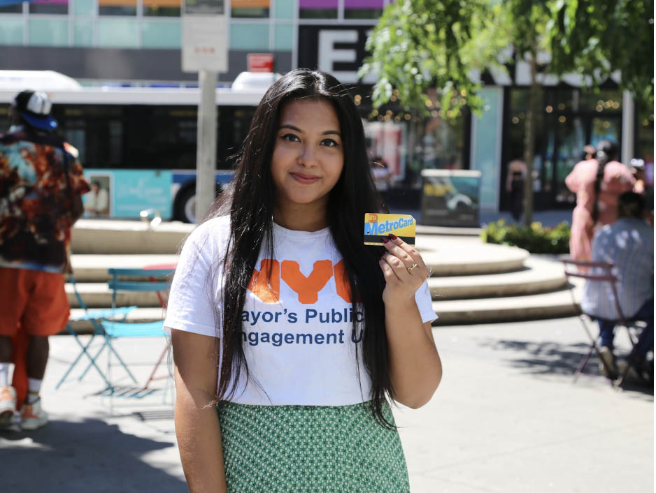 A young woman wearing an NYC Mayor's Public Engagement tee-shirt smiles at the camera and holds up a metrocard