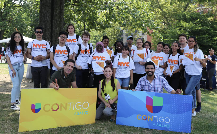 A group of PEU interns all wearing NYC Mayor's Public Engagement tee-shirts and Univision staff  holding 'Contigo A Salvo' signs pose together at the park .