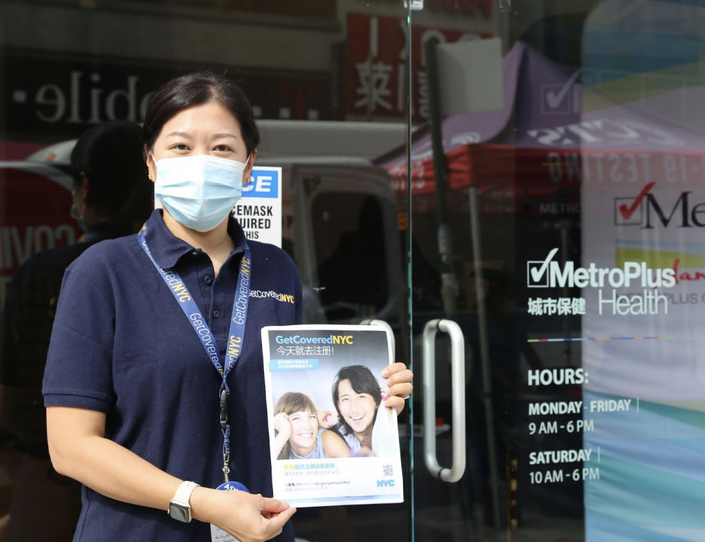 Young woman stands in front of clear doors wearing a mask. She holds a flyer that reads 'GetCoveredNYC'