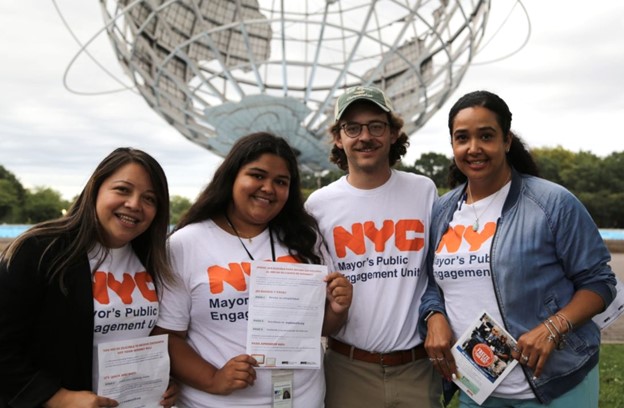 Four PEU staffers stand in together, holding PEU flyers and wearing PEU tee-shirts. They stand in front of the Unisphere, a very large steel statue of the world from the World's Fair in Flushing Meadows–Corona Park.