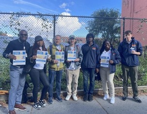 PEU staff stand together on the sidewalk, in front of a community garden, holding up flyers.