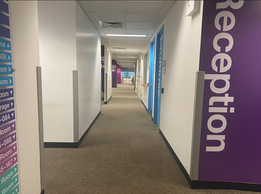 A long pathway with walls and light blue doors.  On the left is a cubicle with computer. On the right there is part of a purple wall with the word Reception painted with white letters.