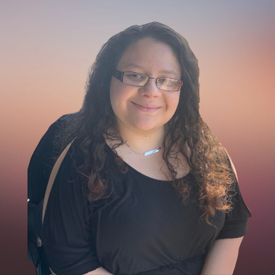 Mandi with long, curly brown hair and glasses smiling at the camera. She is wearing a black open-arm top and a silver necklace with a rectangular pendant. Her background is a gradient of light pink to soft purple. She is seated in a wheelchair, which is slightly visible behind her.