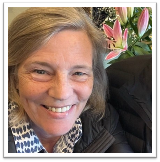 Sara is seated in front of some pink and white flowers and is smiling with grey-brown hair and freckles on her cheeks wearing a white and navy-dotted shirt and a black puffy jacket.