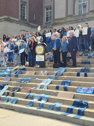 Picture of Staten Island Denim Day Celebration with Richmond County District Attorney Michael McMahon on the steps of Staten Island Borough Hall. A crowd of supporters in denim hold signs and below the podium on the steps are decorated denim jeans and signs