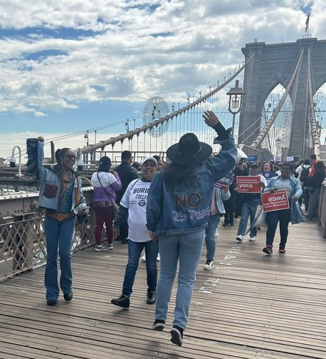 Woman in a dark brimmed hat is facing the crowd, waving her hand to guide them. The back of her denim jacket says No means No and 25 years