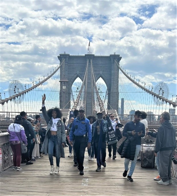 Picture of crowd in denim walking along the Brooklyn Bridge pedestrian walkway. In the front is a woman and man addressing the camera in customized denim outfits.