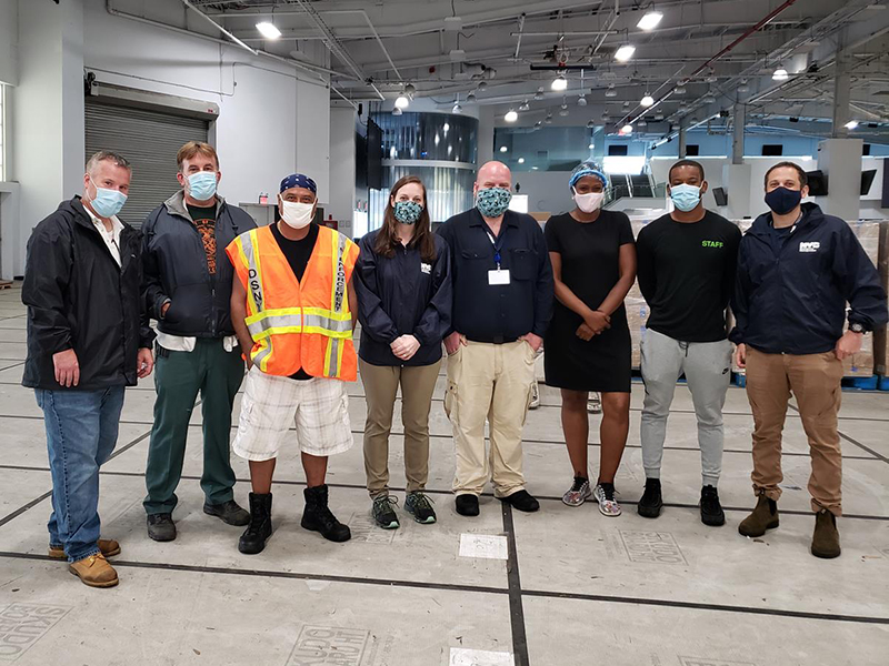 8 paople standing in a warehouse all wearing masks